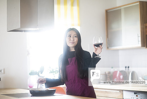 Junge Frau kocht zu Hause und hält ein Glas Rotwein