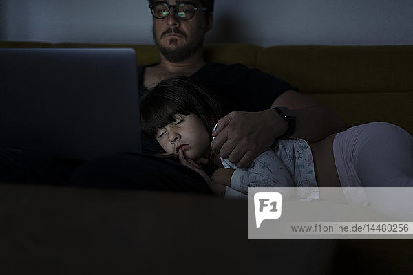 Father using laptop on couch at night with daughter sleeping