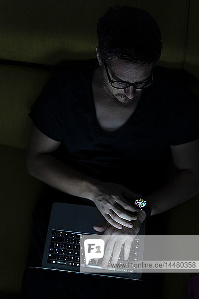 Man sitting on couch using laptop and smartwatch at night