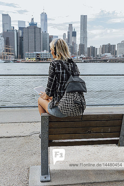 USA  New York City  Brooklyn  junge Frau sitzt am Wasser mit Rucksack  Kopfhörer und Tablett