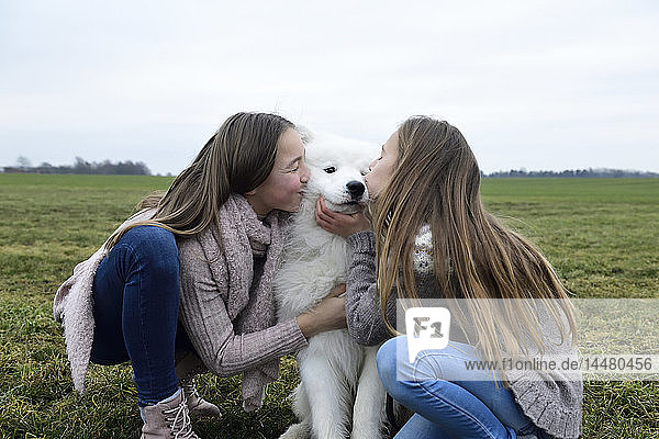 Zwei Mädchen kauern auf einer Wiese und küssen einen Hund