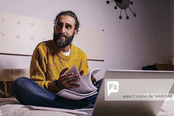 Young man working in bed at home with laptop and documents