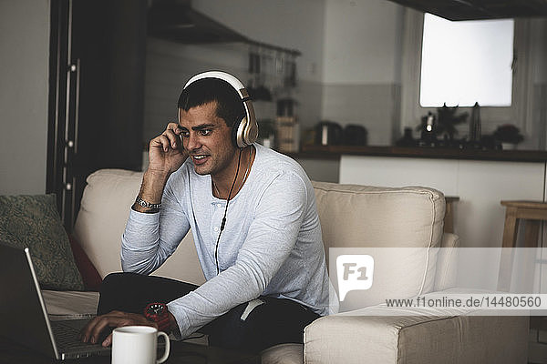 Smiling young man sitting on sofa at home wearing headphones and using laptop