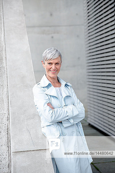 Porträt einer zufriedenen reifen Geschäftsfrau  die in hellblauem Trenchcoat an der Wand lehnt