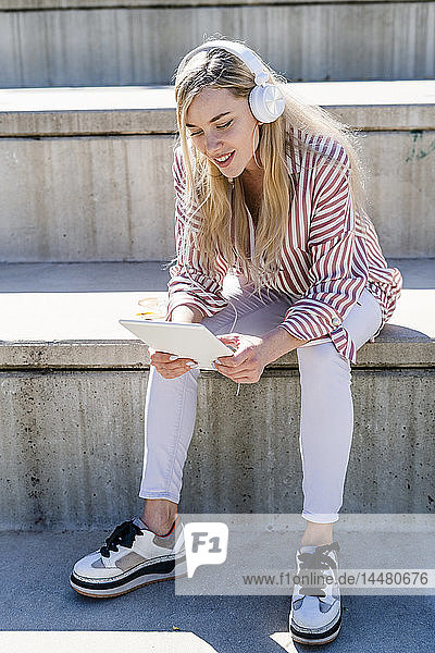 Porträt einer blonden jungen Frau  die auf einer Treppe im Freien sitzt und ein digitales Tablett und Kopfhörer benutzt