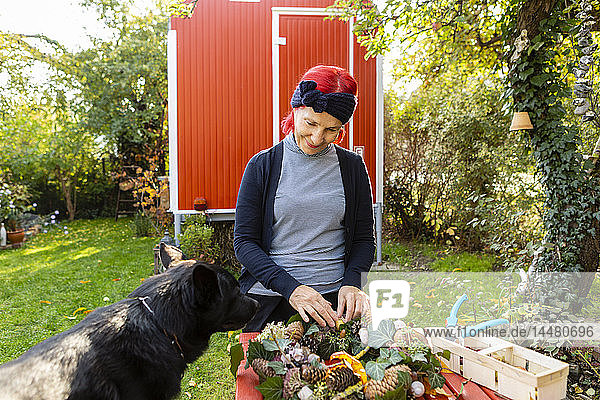 Lächelnde ältere Frau mit rot gefärbtem Haar mit Hund bastelt im Garten an der Herbstdekoration