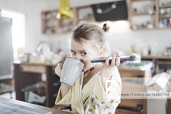 Portrait of little girl with smartphone drinking tea in the kitchen