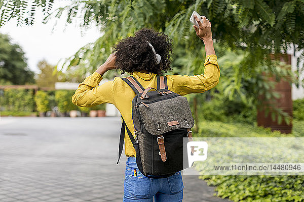 Rückenansicht einer Frau mit Rucksack  die im Freien mit Kopfhörern und Smartphone Musik hört