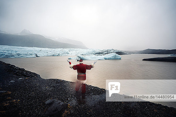 Island  Rückansicht eines als Weihnachtsmann verkleideten Mannes  der an einem Gletscher steht und die Arme hebt