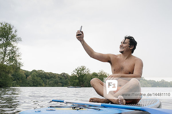 Mann sitzt auf einem SUP-Brett auf einem See und macht ein Selfie