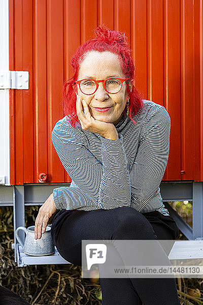 Porträt einer zufriedenen älteren Frau mit rot gefärbten Haaren  die vor einem roten Wohnwagen im Garten sitzt