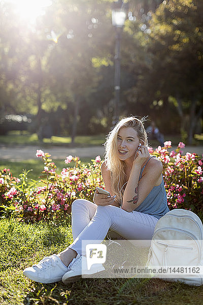 Porträt einer lächelnden jungen Frau  die mit Handy und Ohrstöpseln im Park sitzt