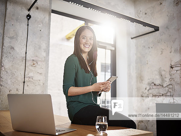 Smiling woman sitting on desk in office listening to music