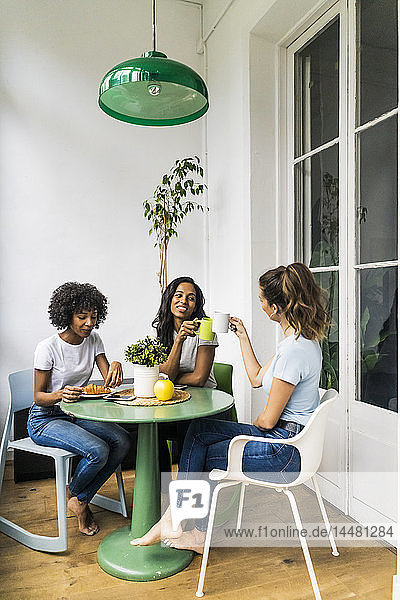 Drei glückliche Frauen sitzen zu Hause am Tisch und trinken gemeinsam Kaffee