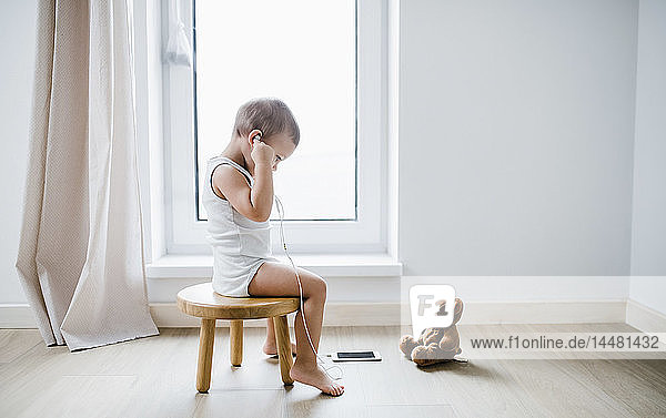Toddler boy sitting on stool at home using smartphone and earphones