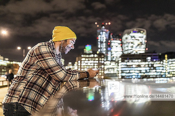 Großbritannien  London  lächelnder Mann lehnt an einem Geländer und schaut auf sein Telefon mit Stadtlichtern im Hintergrund