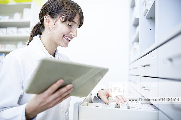 Smiling pharmacist with tablet at cabinet in pharmacy