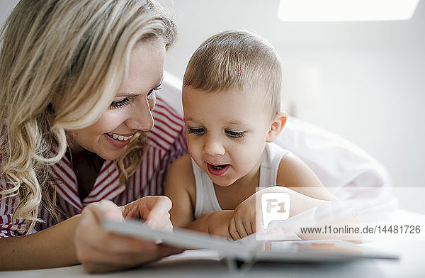 Smiling mother and toddler son lying in bed at home reading a book