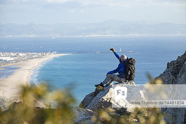 Spanien  Andalusien  Tarifa  Mann auf einer Wanderung an der Küste  der auf einem Felsen sitzt und sich ein Selfie