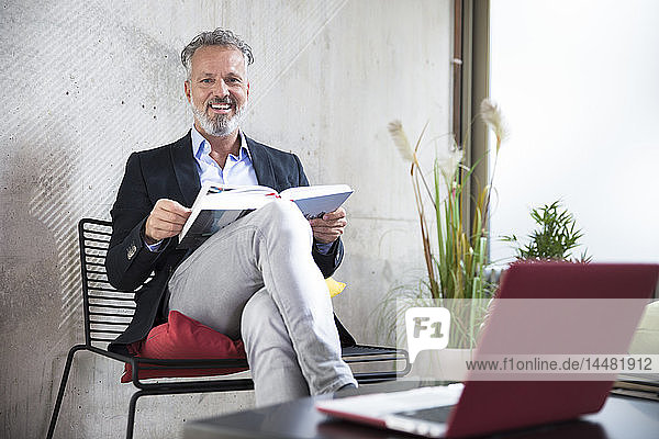 Portrait of happy businessman sitting at concrete wall reading book