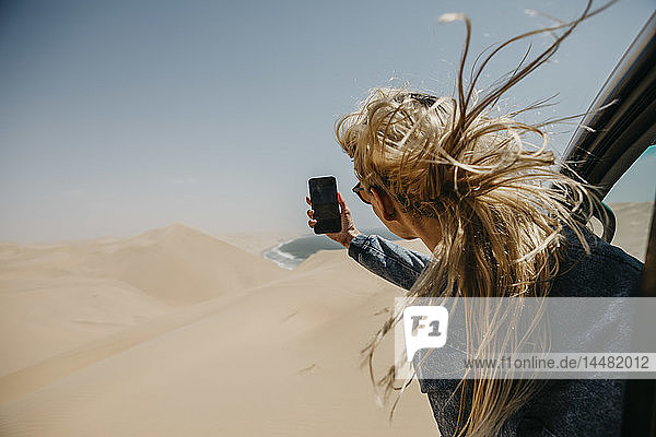 Namibia  Walvis Bay  Namib-Naukluft-Nationalpark  Sandwich Harbour  aus dem Autofenster gelehnte Frau beim Fotografieren mit dem Handy