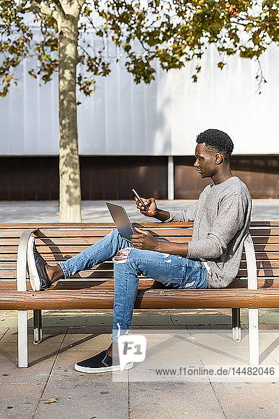 Junger Mann sitzt auf einer Bank in der Stadt und benutzt Laptop und Smartphone