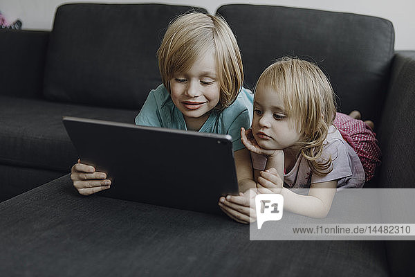 Portrait von Bruder und kleiner Schwester  die zu Hause auf der Couch liegen und ein digitales Tablet benutzen
