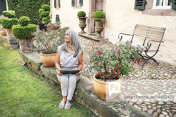 Frau mit langen grauen Haaren sitzt mit Tablette auf der Terrasse eines Landhauses