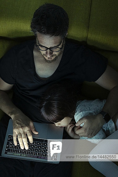 Father using laptop on couch at night with daughter lying next to him