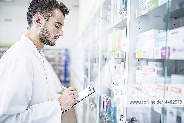 Pharmacist holding clipboard at shelf in pharmacy