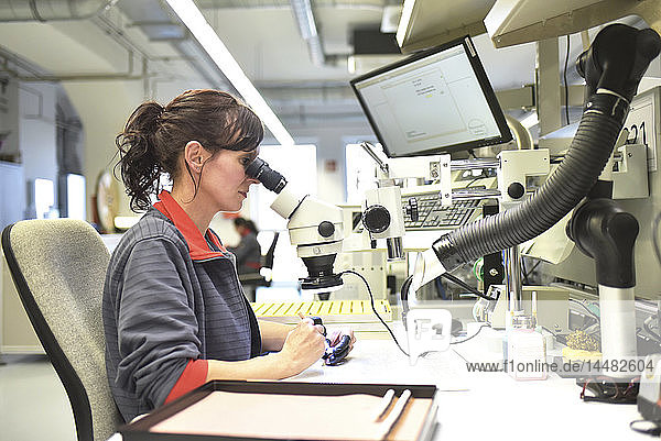 Frau  die ein Mikroskop für die Qualitätskontrolle bei der Herstellung von Leiterplatten für die Elektronikindustrie benutzt