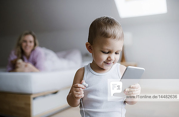 Toddler boy with his mother in bedroom at home using smartphone