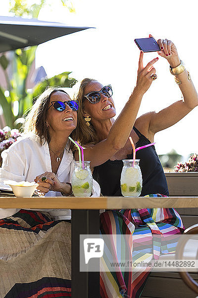 Glückliche Freundinnen  die draußen mit Cocktailgläsern sitzen und ein Selfie trinken