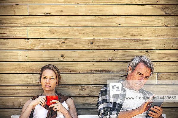 Frau mit Kaffeetasse und Mann mit Smartphone vor einer Blockhütte