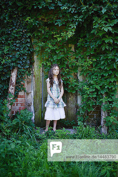 Gelassenes Mädchen im Kleid vor einer rustikalen  mit grünem Efeu bewachsenen Tür stehend