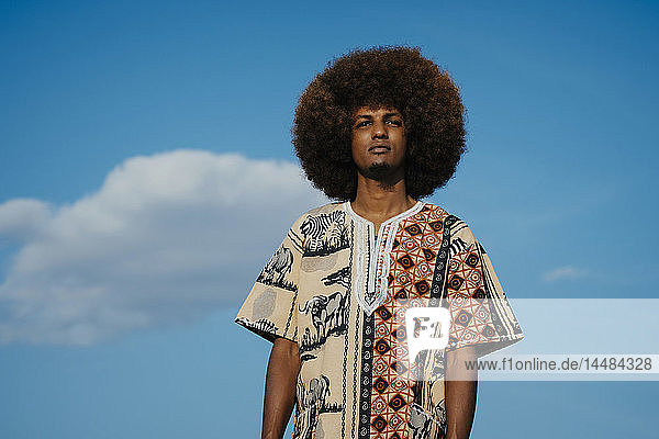 Porträt selbstbewusster junger Mann mit Afro gegen blauen Himmel mit Wolken stehend