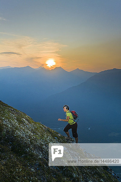 Hiker on the steep hillside of Mt. Alyeska watching the sun set over Girdwood Valley in Southcentral Alaska