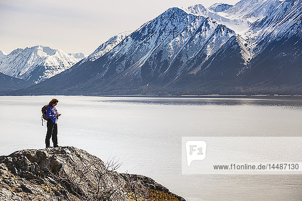 Frau benutzt ein Mobiltelefon  während sie auf einem Felsvorsprung mit Blick auf den Turnagain Arm entlang des Seward Highway steht  Süd-Zentral-Alaska  Frühling