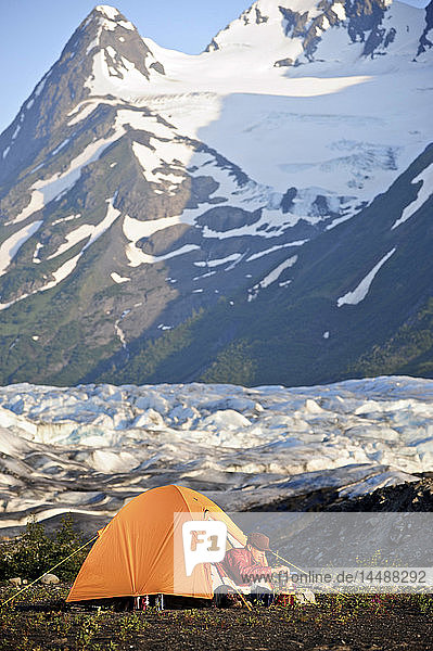 Frau bereitet Essen auf einem Rucksackkocher zu  während sie in ihrem Zelt sitzt  mit dem Spencer Glacier und den Chugach Mountains im Hintergrund  Chugach National Forest  Southcentral Alaska  Sommer