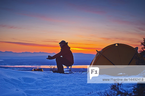 Ein Rucksacktourist wärmt sich die Hände am Lagerfeuer auf einem Campingplatz hoch in den Chugach Mountains mit Blick auf den Kinik Arm und das Cook Inlet mit der Sleeping Lady im Hintergrund  Süd-Zentral-Alaska