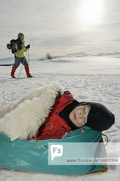 Boy sleeping on sleeping bag with woman walking in the background