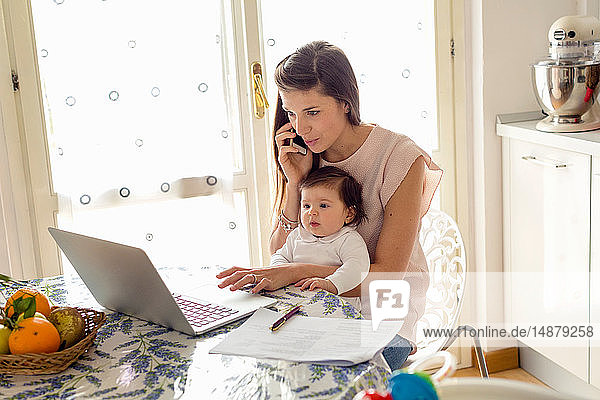 Mother working on laptop with baby girl on lap at home