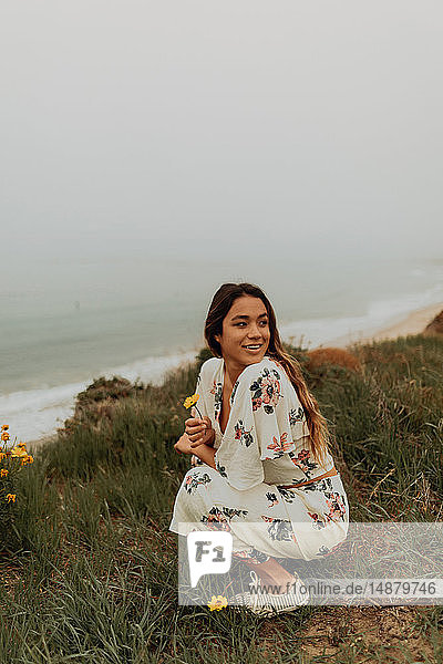 Junge Frau  die an der Küste kauert und Wildblumen hält  Jalama  Kalifornien  USA