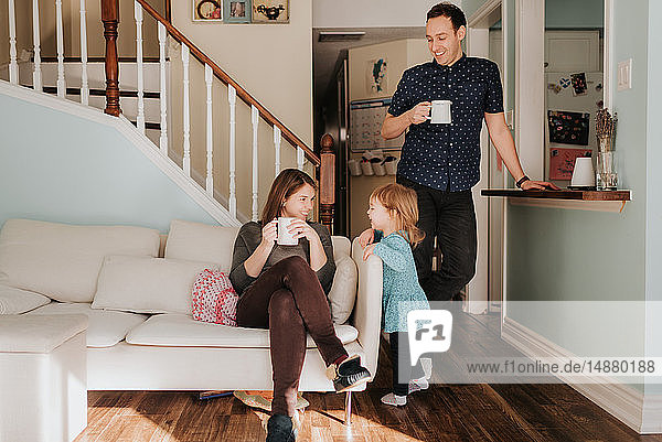 Paar mittlerer Erwachsener  das mit seiner Kleinkind-Tochter eine Tasse Kaffee im Wohnzimmer trinkt