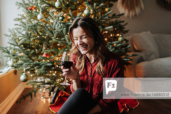 Frau lacht mit Wein neben geschmücktem Weihnachtsbaum