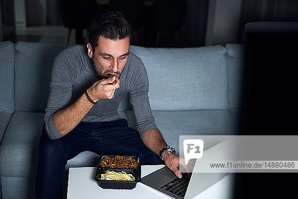 Junger Mann sitzt abends auf dem Sofa  isst zum Mitnehmen und benutzt Laptop