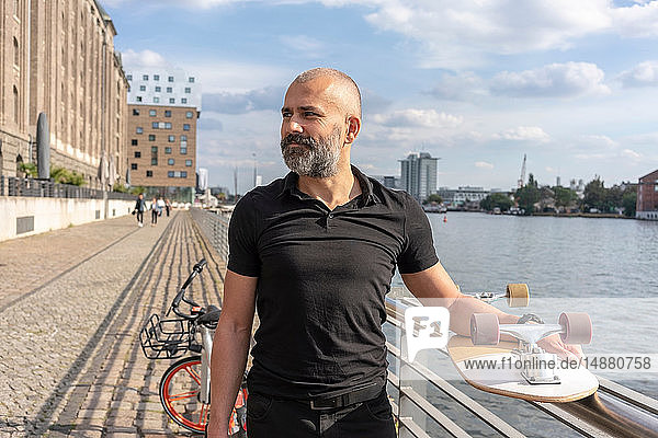 Mann hält Skateboard auf Brücke Fluss und Gebäuden im Hintergrund Berlin Deutschland