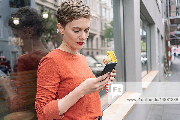Frau hält Eiscreme in der Hand und benutzt ein Smartphone vor dem Geschäft  Köln  Nordrhein-Westfalen  Deutschland