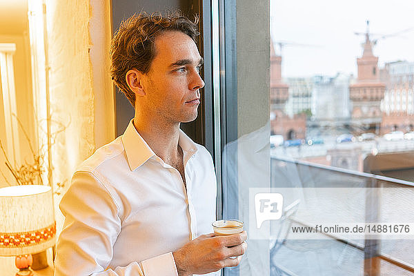 Mid adult businessman looking through office window while taking coffee break