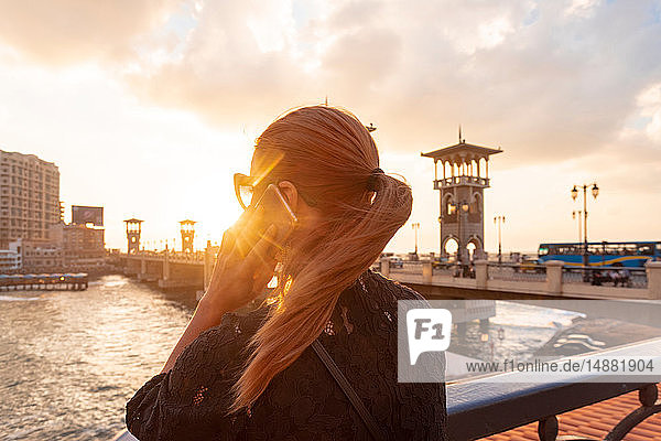 Female tourist with red hair making smartphone call near Stanley bridge at sunset  rear view  Alexandria  Egypt